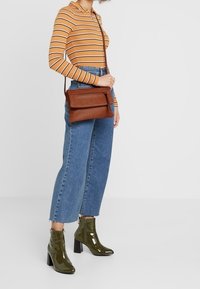 Brun läder axelväska med dragkedjestängning, en framficka och en lång justerbar rem, bärs ihop med blå raka jeans och gröna ankelstövlar.