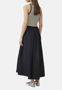 Gestreifter, ärmelloser Oberteil, der in einen schwarzen, bodenlangen Rock mit seitlichen Taschen übergeht. Silberne flache Schuhe vervollständigen das Outfit.
