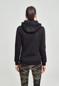 Sudadera negra con bolsillo canguro y capucha con cordón, combinada con leggings de camuflaje que presentan diferentes tonos de verde y patrones negros.