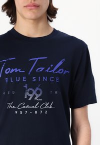 Marinblå bomull T-skjorte med stor hvit og lilla tekst, rund hals og avslappet passform. Teksten inkluderer "Tom Tailor" og "The Casual Club."