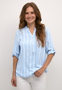 Camisa de botones azul claro con rayas verticales blancas, con cuello de pico y mangas enrolladas, hecha de una tela ligera.