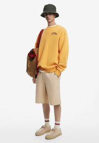 Gul bomulds-sweatshirt med lille grafik, beige shorts og brune sko. Tilbehør inkluderer en brun taske med røde detaljer og en grøn bøttehat.