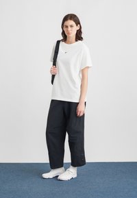 Fiatal nő fehér Nike pólót, laza fekete nadrágot, fehér tornacipőt visel, és egy fekete válltáskát hordoz, miközben kék szőnyegen áll.