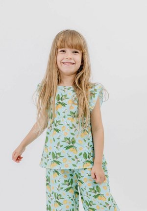 Young girl smiling, wearing light blue outfit with yellow lemon and green leaf print, standing against a plain white background.