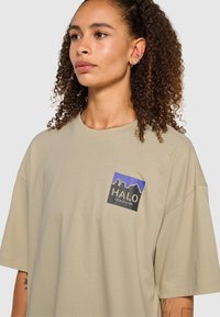 Beige bomullströja med lös passform, med en fyrkantig grafik i blått och svart med texten "HALO" och en stadsbilddesign på vänster bröst.