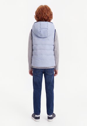 Kind mit rotem Haar steht mit dem Rücken zur Kamera, trägt eine hellblaue wattierte Weste mit Kapuze, ein graues Langarmshirt, blaue Jeans und dunkle Turnschuhe.