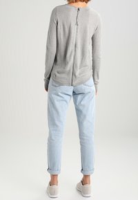 Haut gris à manches longues avec détail de fermeture éclair centrale, associé à un jean bleu clair aux revers retroussés et des chaussures grises, vu de dos.