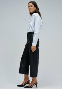 Mujer vistiendo una camisa blanca de botones, un suéter negro de cuello alto, pantalones negros de pierna ancha y recortados, y tacones negros de tipo slingback, de pie de lado.