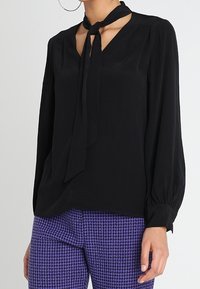 Blusa nera con scollatura a V e dettaglio con fiocco al colletto, dotata di maniche lunghe e una texture liscia. Abbinata a pantaloni a quadri viola.