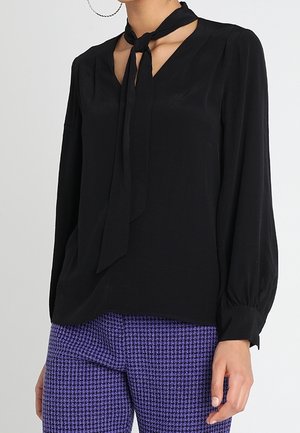 Blusa nera con scollatura a V e dettaglio con fiocco al colletto, dotata di maniche lunghe e una texture liscia. Abbinata a pantaloni a quadri viola.