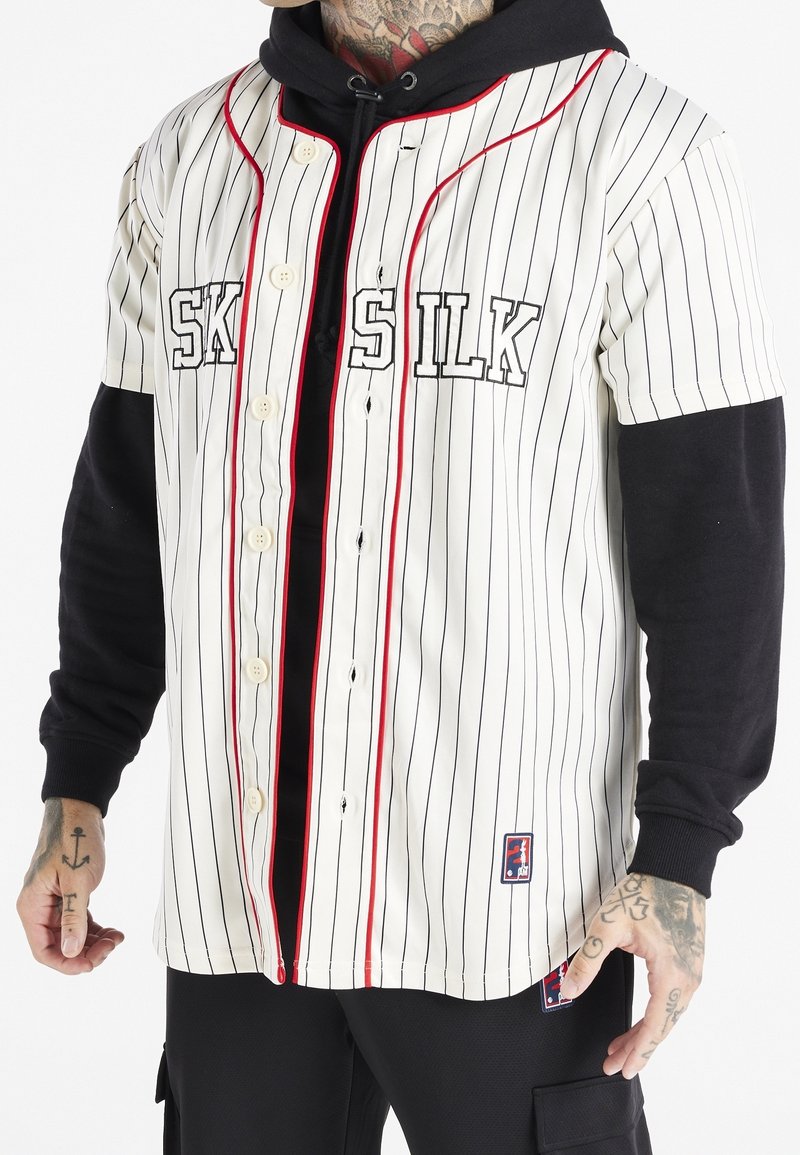 Homme portant un maillot blanc à fines rayures avec boutons, bordure rouge et texte « SILK » sur un sweat à capuche noir, montrant ses mains et son cou tatoués.