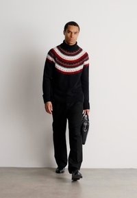 Homme marchant avec un pantalon noir et un pull col roulé noir, orné d'un empiècement rouge et blanc à motifs, tenant un sac en cuir noir dans la main droite.