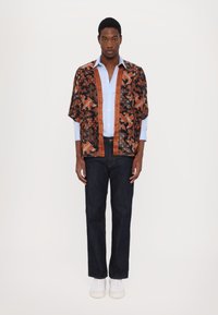 Zwarte kimono-stijl jas met een patroon en oranje accenten, gedragen over een lichtblauw shirt, gecombineerd met donkere jeans en witte sneakers.