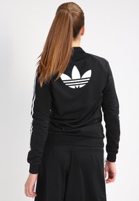 Kvinna med lång brun hästsvans iförd svart Adidas-jacka och byxor, vänd bort, visar det stora vita Trefoil-logotypen på jackans baksida.