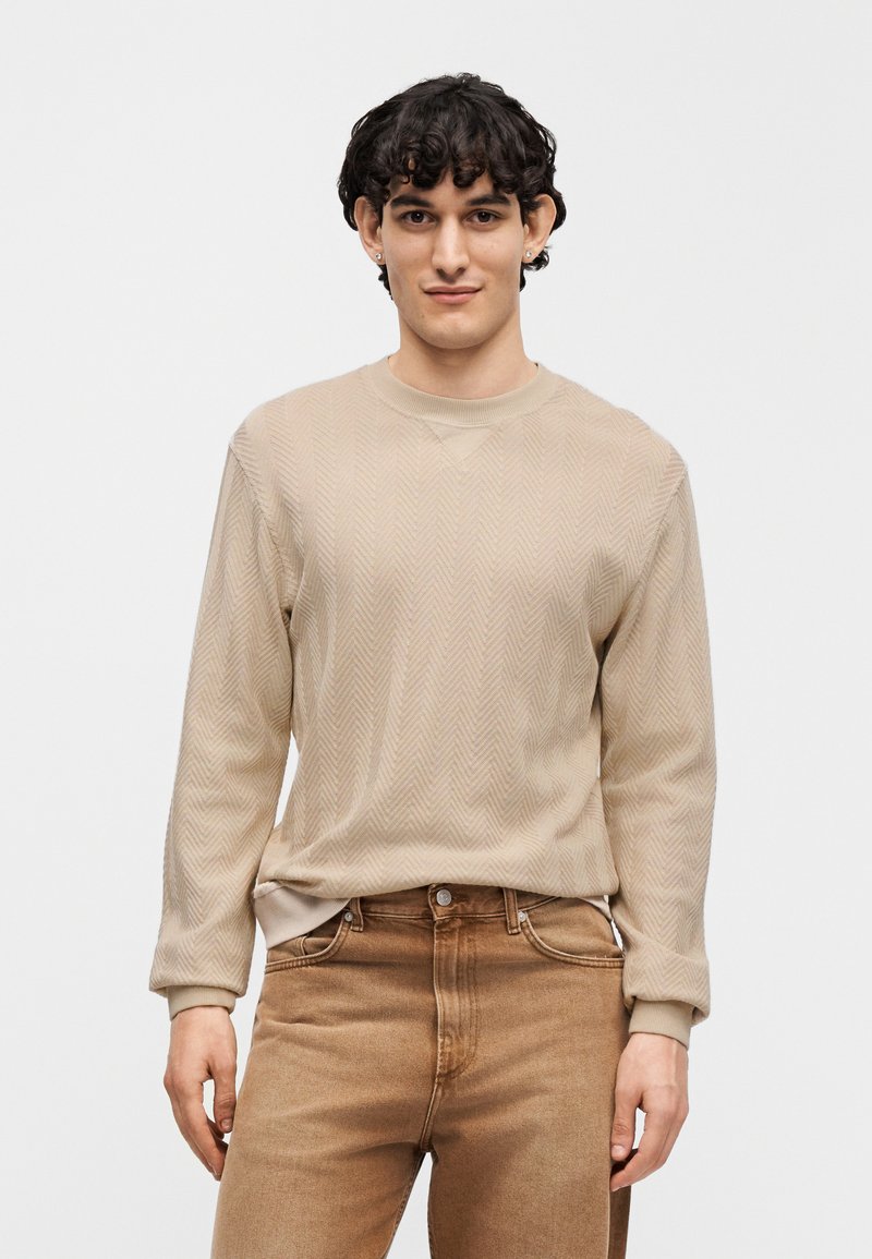 Pull beige en chevrons à manches longues et encolure ronde, associé à un jean marron clair. Le tissu semble doux et texturé.