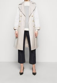 Beige trenchcoat med dubbelknäppta guldknappar, stor krage, långa ärmar och vita ärmar. Matchas med grå tröja och mörka jeans.