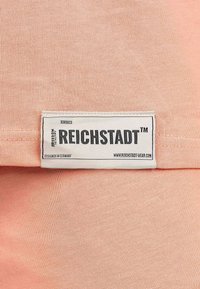 Tessuto color pesca chiaro con una trama liscia che mostra un'etichetta con il marchio, caratterizzata da testo nero su sfondo bianco, cucita su un lato.