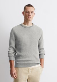 Jasnoszary sweter z dzianiny o fakturowanym wzorze, okrągłym dekolcie i ściągaczach przy mankietach. Model zestawiony z jasnymi spodniami, stoi na tle białym.