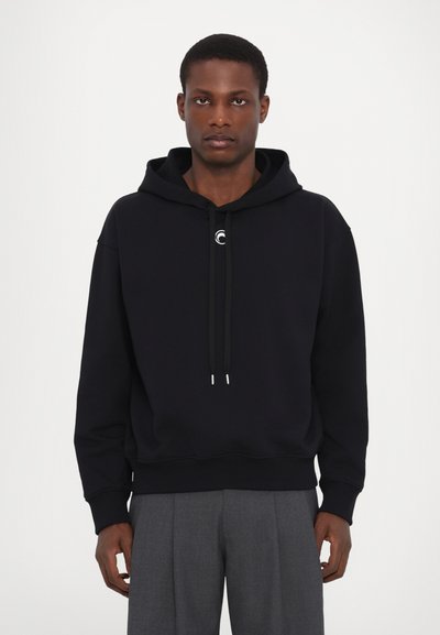 Hanorac negru cu șnururi, cu un logo circular pe piept, asortat cu pantaloni gri plisati și un design minimalist.