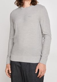 Man som bär en ljusgrå tröja med rund halsringning och "BOSS"-logotyp på bröstet, tillsammans med mörkgrå prickiga byxor, som står mot en enfärgad bakgrund.