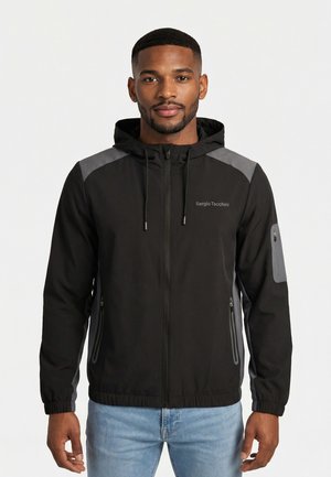 Uomo che indossa una giacca nera e grigia con cappuccio e zip, tasche con zip e jeans azzurri, rivolto in avanti su sfondo bianco.