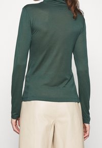 Eine Person trägt ein enges, dunkelgrünes Langarm-Turtleneck-Oberteil und helle beigefarbene Hosen, von hinten vor einem einfarbigen Hintergrund gezeigt.