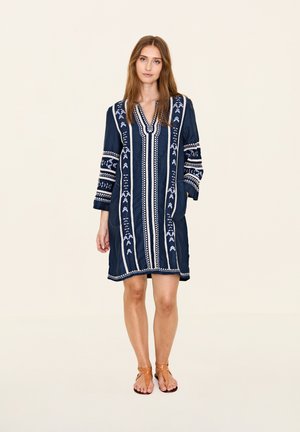 Marineblaues, knielanges Kleid mit filigraner weißer Stickerei, V-Ausschnitt und drei Viertel-Ärmeln. Verfügt über zwei seitliche Taschen und eine lockere Passform.