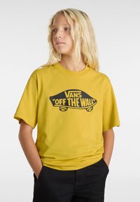 Gul bomullströja med en svart "Vans 'Off the Wall'" logotyp. Korta ärmar, rundad halsringning, avslappnad passform. Bärs med svarta byxor.