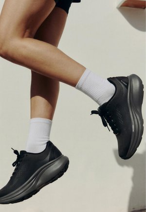 Jambes portant des chaussures de course noires et des chaussettes blanches, capturées en l'air sur un fond clair uni.