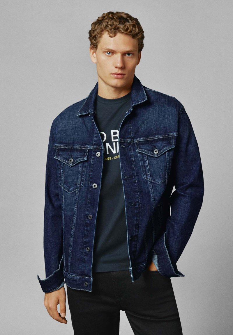 Giubbotto di denim blu scuro, con design classico caratterizzato da bottoni automatici e due tasche sul petto. Indossato sopra una maglietta nera, posato su uno sfondo neutro.