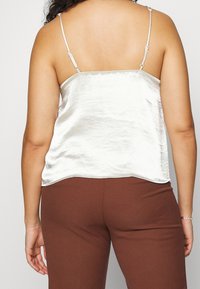 Silver satin camisole with thin adjustable straps, a smooth texture, and a straight hemline, paired with brown fitted pants.