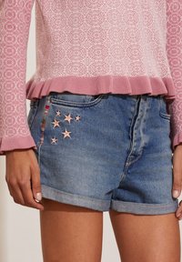 Shorts en denim avec des ourlets roulés, ornés d'étoiles roses brodées sur la poche. Associés à un pull en tricot à motifs rose clair.