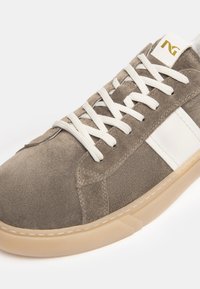 Zapatillas de ante marrón con acentos blancos, suela de goma y cordones planos blancos. Presenta un logo en la lengüeta y detalles de costura reforzada.