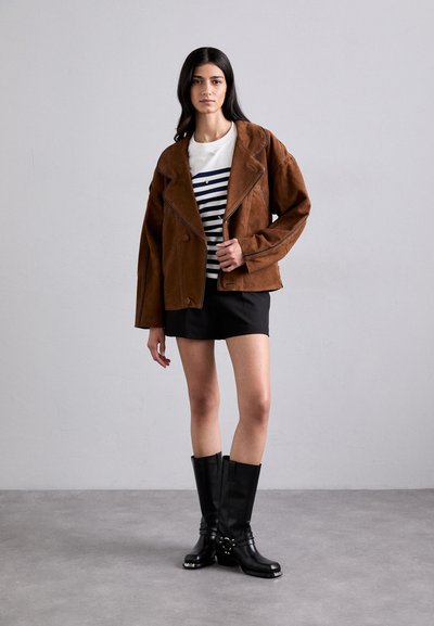 Giacca in suede marrone stile motociclista con grande colletto, strisce nere su una camicia bianca, mini gonna nera e stivali neri al ginocchio.