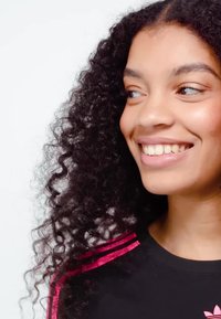 Mladá žena s kudrnatými vlasy se usmívá a dívá se stranou, má na sobě černý top s růžovými pruhy na rameni.