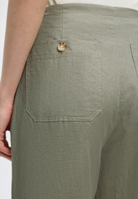 Vedere din spate a pantalonilor de in într-o nuanță deschisă de măsliniu, cu un singur buzunar cu nasture la spate, iar partea din mâna purtătorului și o bluză albă sunt vizibile.