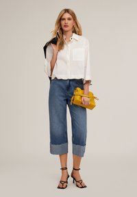 Camicia bianca corta con tasca, jeans azzurri a gamba larga con orli sfrangiati e una borsa gialla. Tacchi neri con cinturini completano l'outfit.