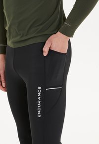 Svarta träningsleggings med en sidficka, vitt 'ENDURANCE'-logotyp och reflekterande detaljer. Materialet verkar vara stretchigt och åtsittande.