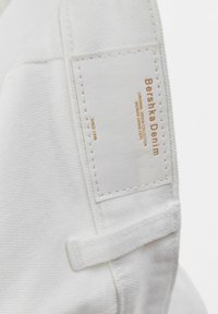 Tissu en denim blanc avec une surface texturée et une étiquette cousue affichant "Bershka Denim" en or. L'étiquette indique "Collection Denim Originale".