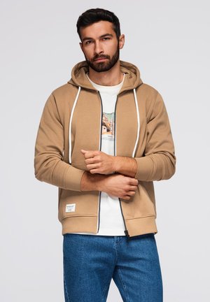 Beige zip-hoodie med hvide snore, ribbede manchetter og kant. Bæres over en hvid T-shirt med grafisk print. Blå denim jeans.
