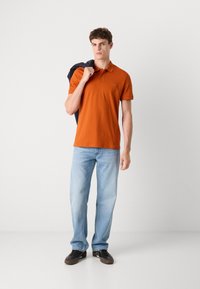 Oransje polo skjorte med kort krage og et merke på brystet, parret med lyseblå jeans og mørke sneakers. Modellen holder en marineblå jakke.