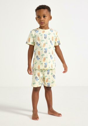Dreng står barfodet iført matchende cremefarvede shorts og t-shirt med farverige hunde- og palmetræsprint på hvid baggrund.