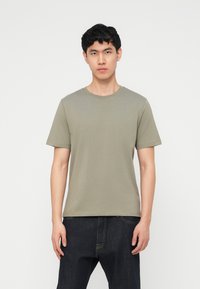 Νέος άνδρας με κοντά μαύρα μαλλιά που φοράει μια κοντομάνικη πράσινη λαδί μπλούζα και μαύρα τζιν, μπροστά σε λευκό απλό φόντο.