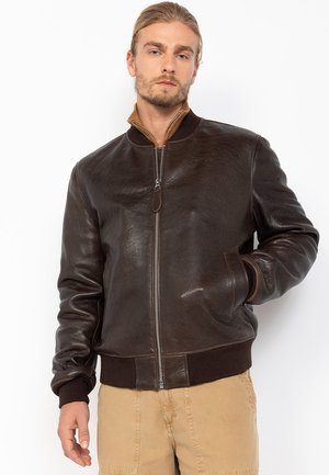 Homme aux cheveux blonds et à la barbe, portant une veste bomber en cuir marron foncé et un pantalon beige, se tenant avec une main dans la poche.