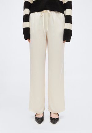 Osoba oblečená v voľných krémových nohaviciach so šnúrkou v páse, čierno-bielom pruhovanom svetri s dlhým rukávom a čiernych špicatých topánkach.