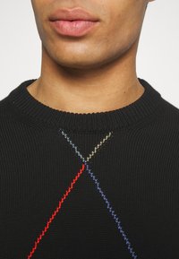 Černý pletený svetr s kulatým výstřihem. Obsahuje barevnou výšivku v červené, modré, zelené a žluté, která tvoří diagonální čáry přes hrudník.