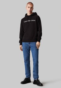 Sudadera con capucha negra y logo blanco de "Calvin Klein Jeans", combinada con jeans de mezclilla azul y zapatos negros de cordones, de pie frente a un fondo gris.
