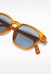 Orange, transparent solglasögon med fyrkantiga bågar och blåtonade linser. Har "GUESS"-logotypen på den högra skalmen och guldaccenter.
