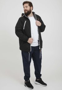 Czarna kurtka przeciwdeszczowa z białym zamkiem błyskawicznym i sznurkami, z kapturem i brązowymi akcentami. Połączona z granatowymi spodniami dresowymi i czarnymi sneakersami.