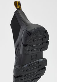 Černá obuv s texturovanou gumovou podrážkou, se hladkým svrškem a poutkem s žlutým označením. Design je zjednodušený a moderní.